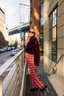 Woman in red fur coat and patterned pants standing on a balcony with a cityscape background