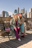 Two women sitting on a bench with a city skyline in the background
