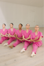 Five dancers in pink pajama sets sitting on a wooden floor.