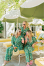 Three women in matching green pajama sets with floral patterns sitting under green umbrellas in a sunlit outdoor setting.