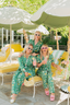 Three women in matching green pajama sets with floral patterns are sitting on a yellow chair under white umbrellas in a sunlit outdoor setting.