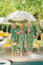 Three women in matching green floral pajamas standing by a pool with yellow chairs and an umbrella.