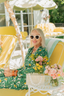 Woman in a green patterned pajamas sitting in a yellow chair with flowers and sunglasses, outdoor setting.