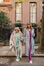 Two women walking on a sidewalk in an urban setting, wearing stylish outfits and holding coffee cups.