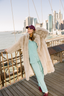 Woman in a fur coat and light blue outfit standing on a bridge with a city skyline in the background