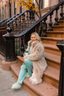 Woman in a fur coat sitting on brownstone steps holding a coffee cup.