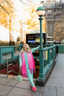 Woman in colorful outfit standing on a city street with a green lamp post and subway entrance in the background.