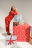 Woman in red pajama set with gift-wrapping theme, leaning against wrapped gifts.