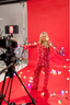 Woman standing on a set with a red backdrop in a matching red pajama set with white bows holding a string of Christmas lights. 